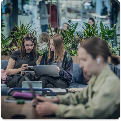 Trzy osoby uczącę się, dwie skupione na tablecie i laptopie, trzecia rozmyta na pierwszym planie korzysta z laptopa. W tle zielone rośliny.